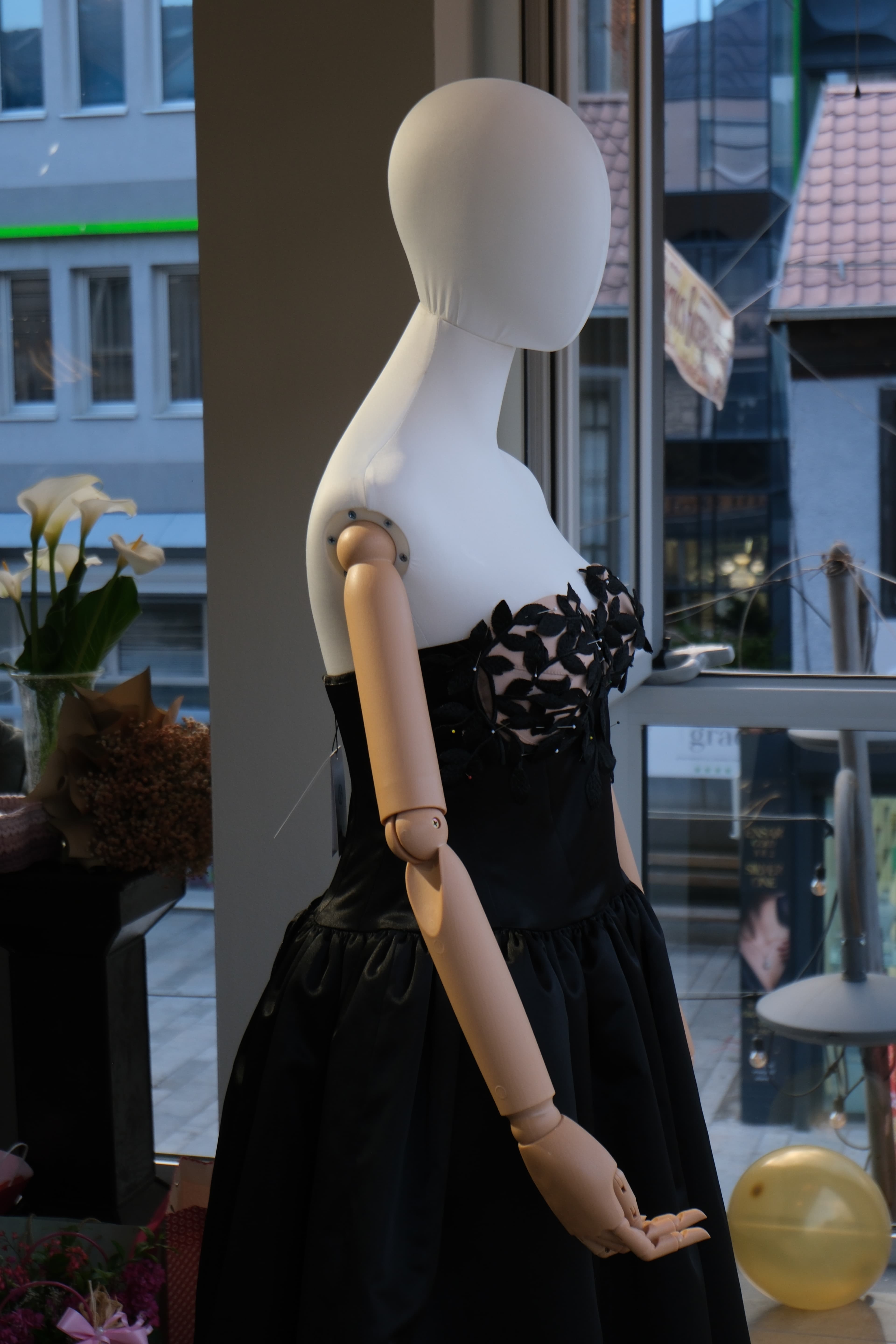 Mannequin wearing the black leaf-appliqué gown at the RABEL opening, calla lilies in the foreground and a gold balloon above
